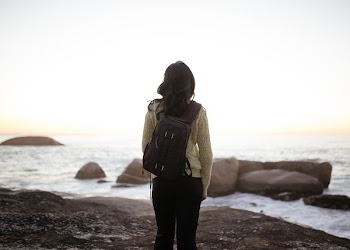 Woman Wearing White Long-sleeved Top and Black Pants Carrying Black Backpack While Standing on Shore during Adytime