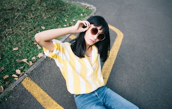 woman wearing yellow and white striped collared shirt holding round-shaped brown sunglasses sitting on gray concrete pavement during daytime