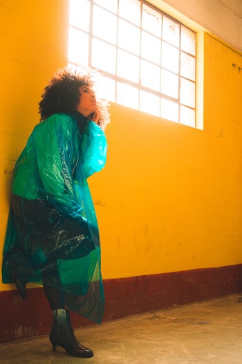 Women's Green Rain Coat Leaning on Yellow Painted Wall
