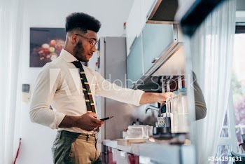 Young businessman holding smartphone while making coffee at home