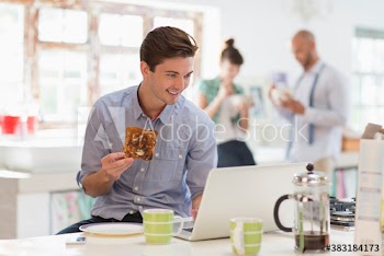 Young man eating toast at laptop in morning kitchen