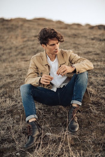Young Man in Blue Jeans and Beige Jacket Sitting on Ground in Empty Fields