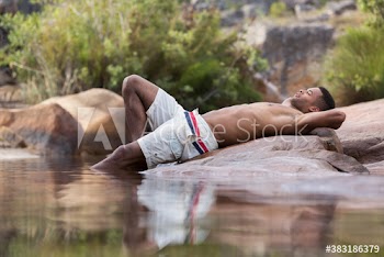 Young man in swim trunks relaxing on rocks at lakeside