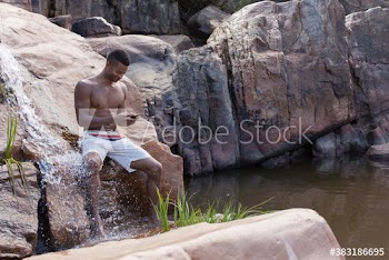 Young man in swim trunks using smart phone on rocks with waterfall