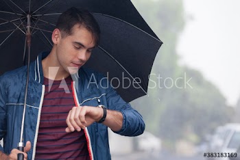 Young man with umbrella checking time on wristwatch in rain