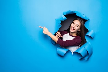 Young pretty woman pointed side while looking through blue hole in paper wall. Free Photo