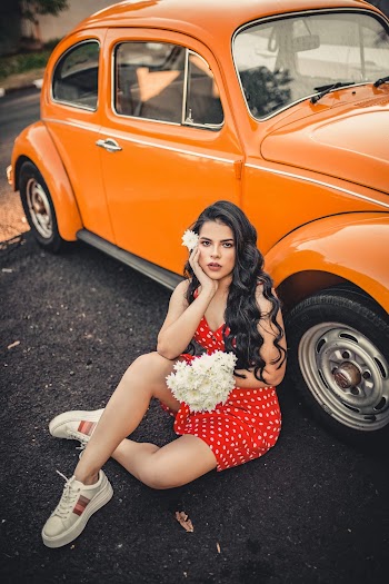 Young Woman In An Orange Polka Dot Dress Holding A Bunch Of White Daisies While Sitting On Concrete Pavement Close To A Volkwagon Beetle